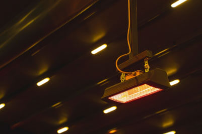 Radiant heating unit suspended from the ceiling inside a large UK factory, showing glowing heating tubes within a metal housing above blue storage racks and industrial shelving.