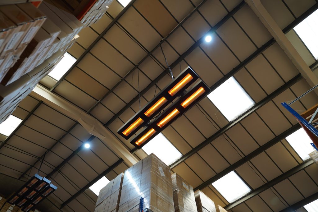 Radiant heating unit suspended from the ceiling inside a large UK factory, showing glowing heating tubes within a metal housing above blue storage racks and industrial shelving.
